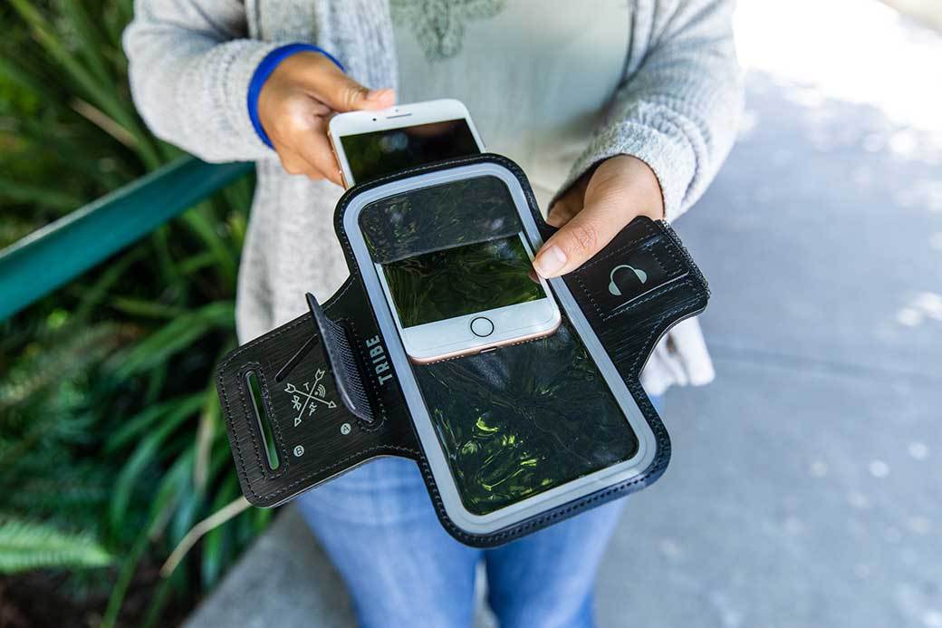 A person in a gray top and light jeans stands on a sun-dappled sidewalk as they take a smartphone out of a fitness armband.